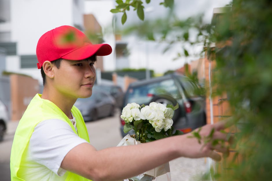 A young male flower delivery courier with light skin and dark hair, wearing a red cap and a high-visibility yellow vest over a white T-shirt, is placing a bouquet of fresh white hydrangeas against a g