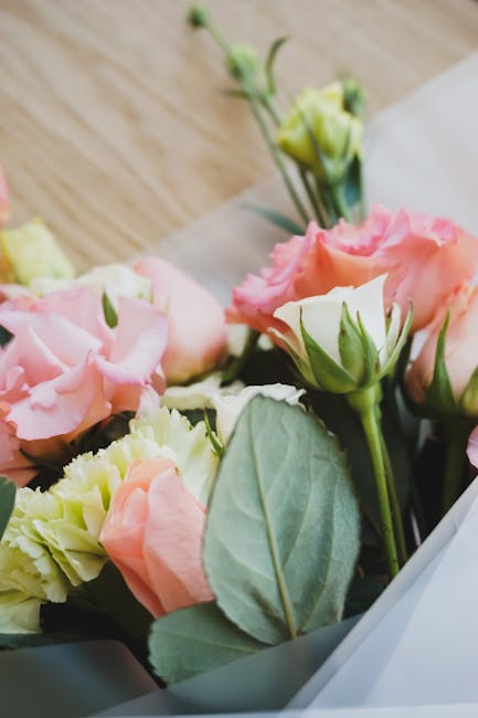 A close-up view of a floral arrangement featuring pale pink and cream lisianthus, accompanied by green foliage with smooth, oval-shaped leaves. The flowers have delicate, ruffled petals with a soft te