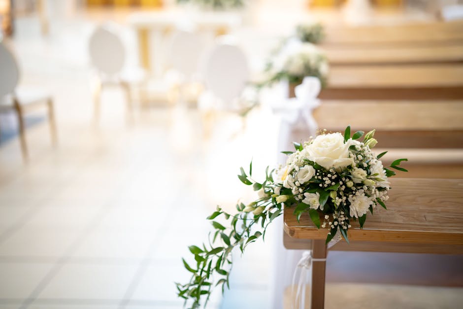 A bouquet of fresh white flowers, including roses, baby's breath, and greenery, resting on a wooden pew in a well-lit indoor setting. The arrangement is delicately placed, with the flowers arranged in