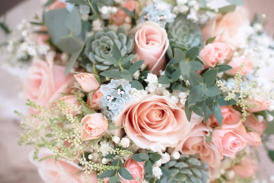 A close-up view of a floral bouquet featuring large, soft pink roses with tightly packed petals, complemented by smaller pink spray roses. The arrangement also includes clusters of white baby's breath