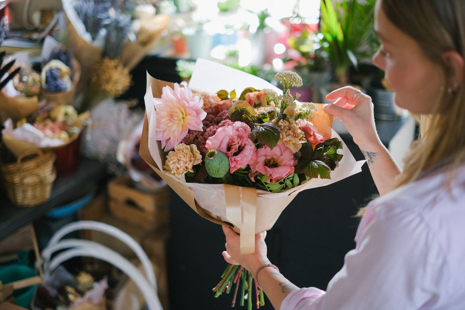 A woman holding a bouquet of fresh flowers in a florist shop. The bouquet features large pink dahlias, pale pink lisianthus, small beige carnations, and green foliage. The flowers are arranged in a be